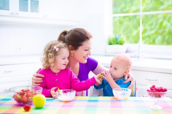 Happy young family, mother with two children, adorable toddler girl and funny baby boy having healthy breakfast eating fruit and dairy, sitting in a white sunny kitchen with window